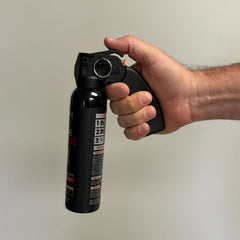 A hand grips the handle of a POM Home Defense 9.2oz Pepper Spray by POM, with the index finger on the trigger, against a plain light background.