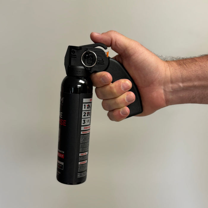 A hand grips the handle of a POM Home Defense 9.2oz Pepper Spray by POM, with the index finger on the trigger, against a plain light background.
